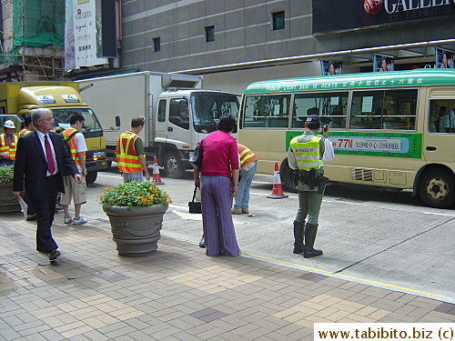 HK policeman. Minibus in the background. Wait a minute! Is HE on the cell phone too?  No, it looks like a walkie talkie. Whew!