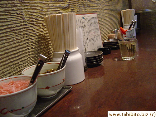The counter top is filled with utensils, condiment and all-you-can-eat pickled ginger and veggies